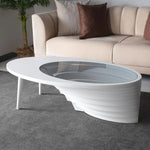 An oval-shaped coffee table with a layered walnut base and a tempered glass top, placed in a room with a sofa and a plant in the background.