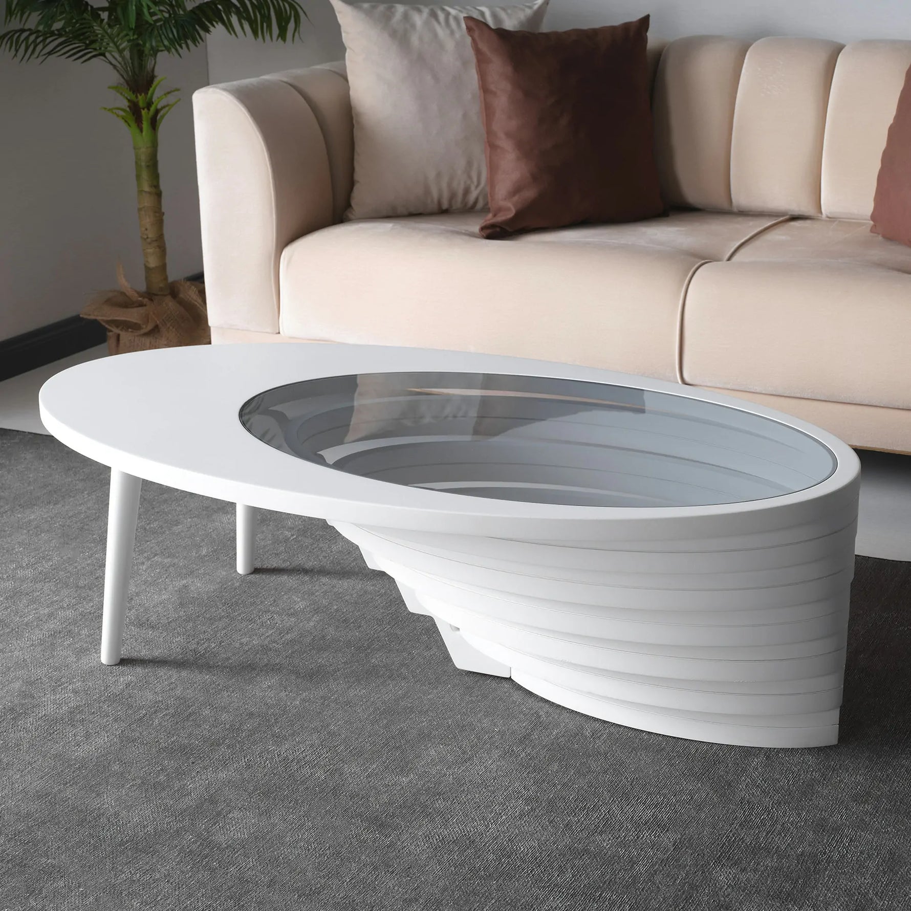 An oval-shaped coffee table with a layered walnut base and a tempered glass top, placed in a room with a sofa and a plant in the background.
