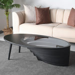 An oval-shaped coffee table with a layered walnut base and a tempered glass top, placed in a room with a sofa and a plant in the background.