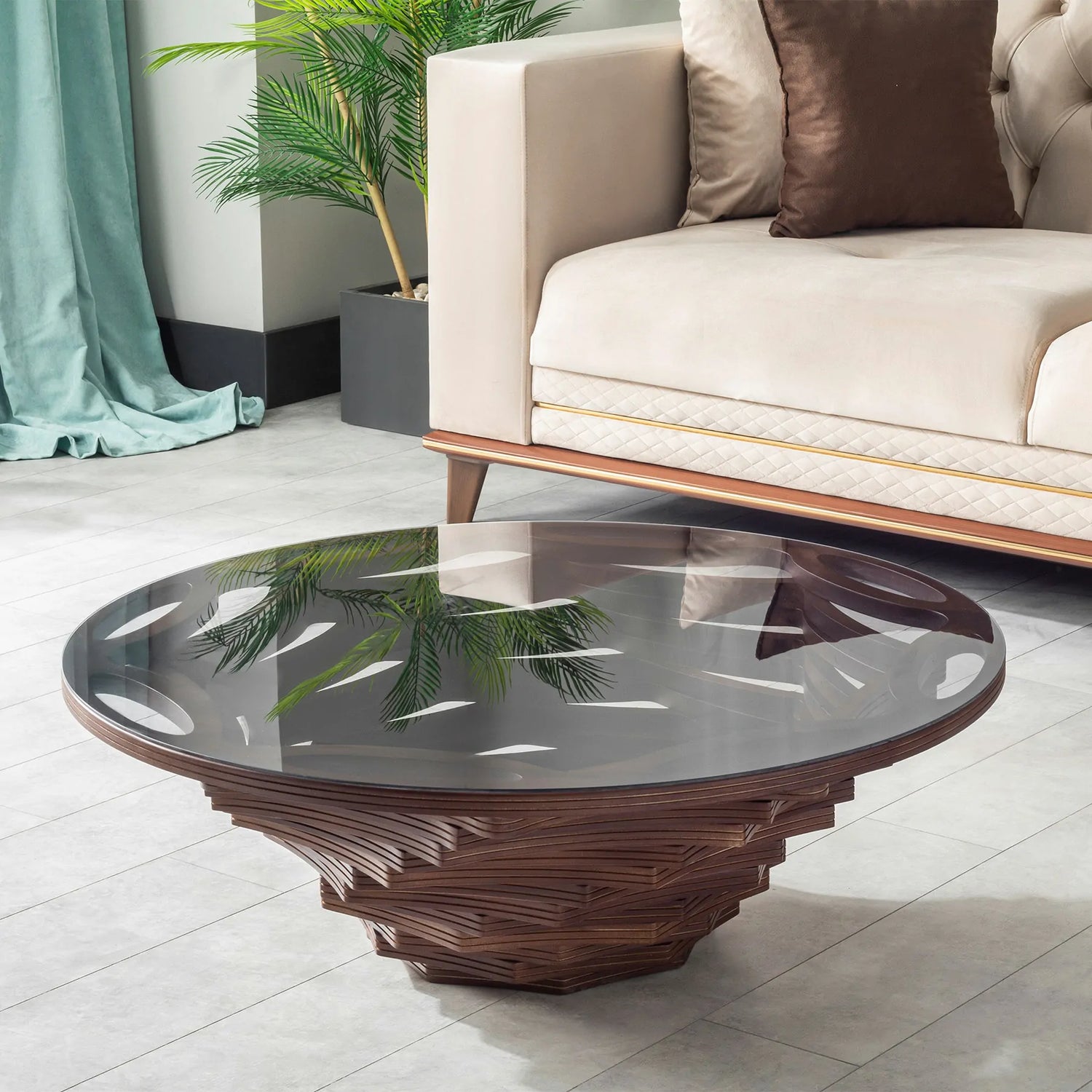A round coffee table with a glass top and a sculptural wooden base, featuring layered walnut accents, placed in a room with a sofa and a plant in the background.