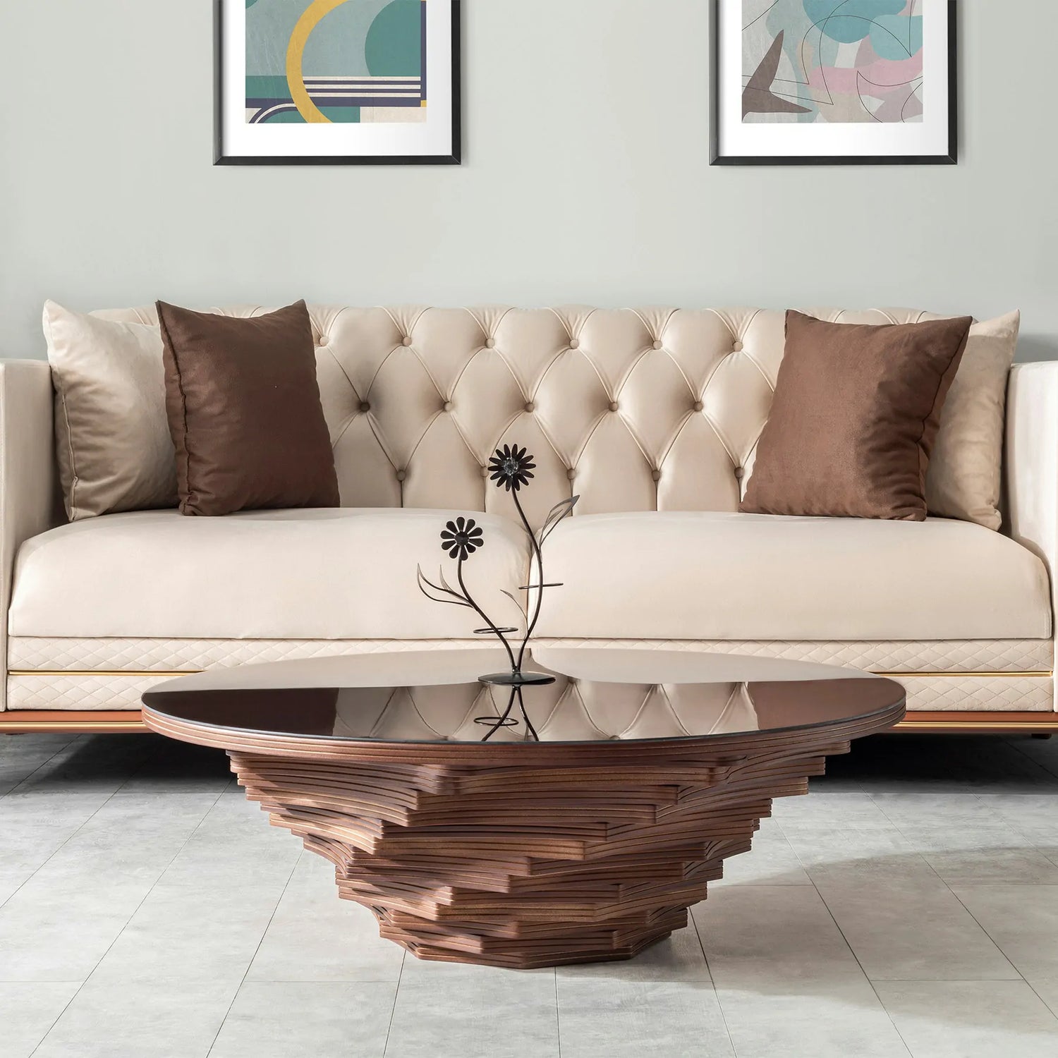 A round coffee table with a glass top and a sculptural wooden base, featuring layered walnut accents, placed in a room with a sofa and a plant in the background.