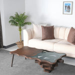 A stylish coffee table with a unique layered design, featuring wood and glass elements, placed in a living room setting with a white sofa and decorative pillows.