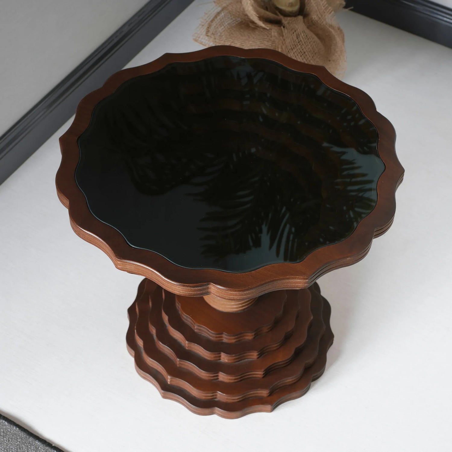 A round wooden side table with a scalloped edge design and a dark glass top, placed on a white floor.