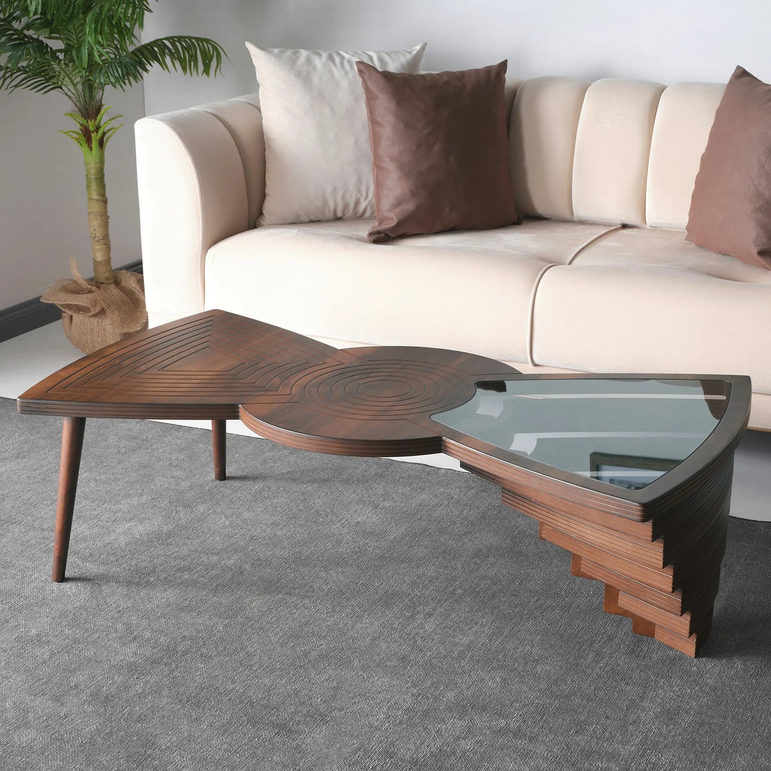 Asymmetrical layered coffee table with wood and glass sections, positioned in a living room setting with a sofa and cushions in the background.