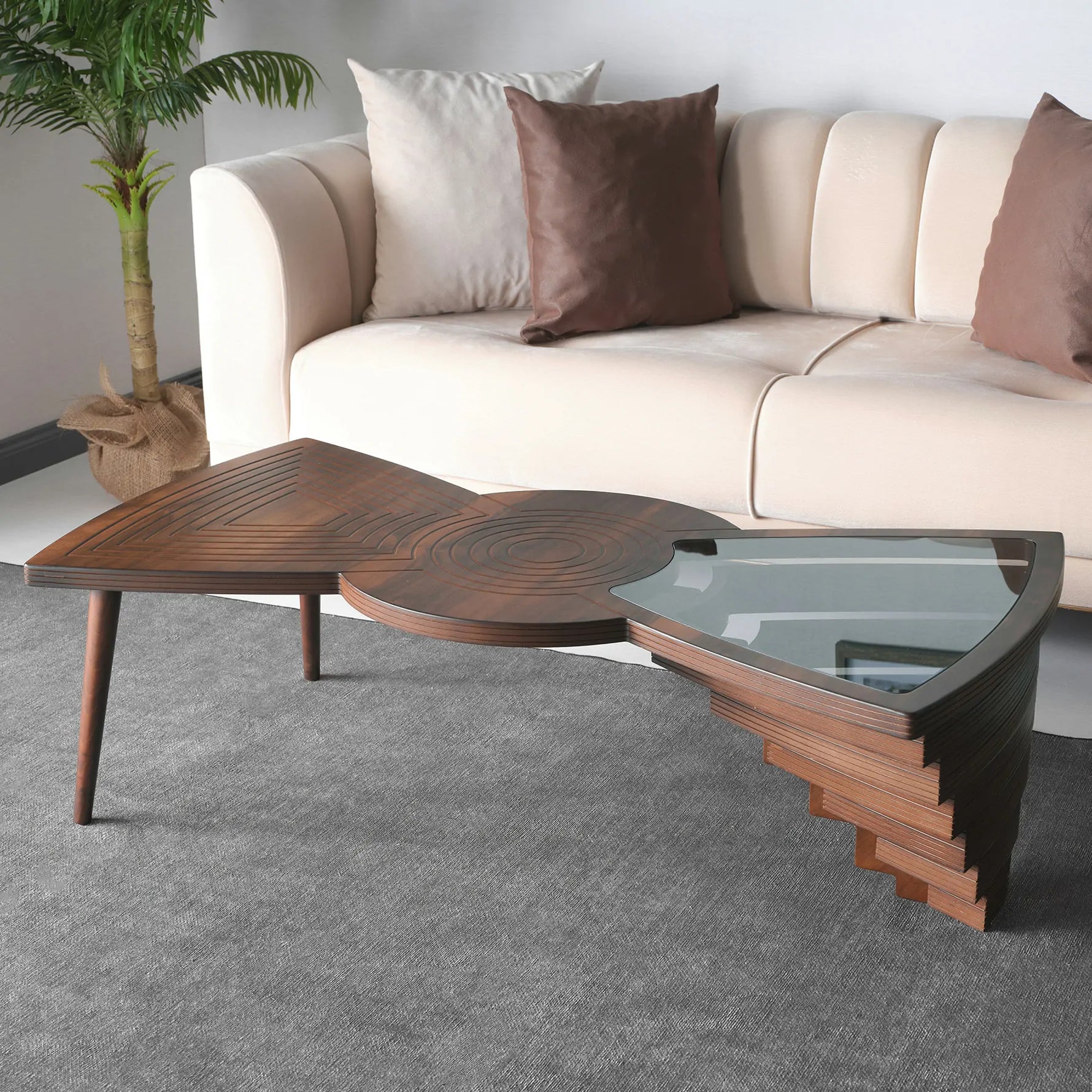 Asymmetrical layered coffee table with wood and glass sections, positioned in a living room setting with a sofa and cushions in the background.
